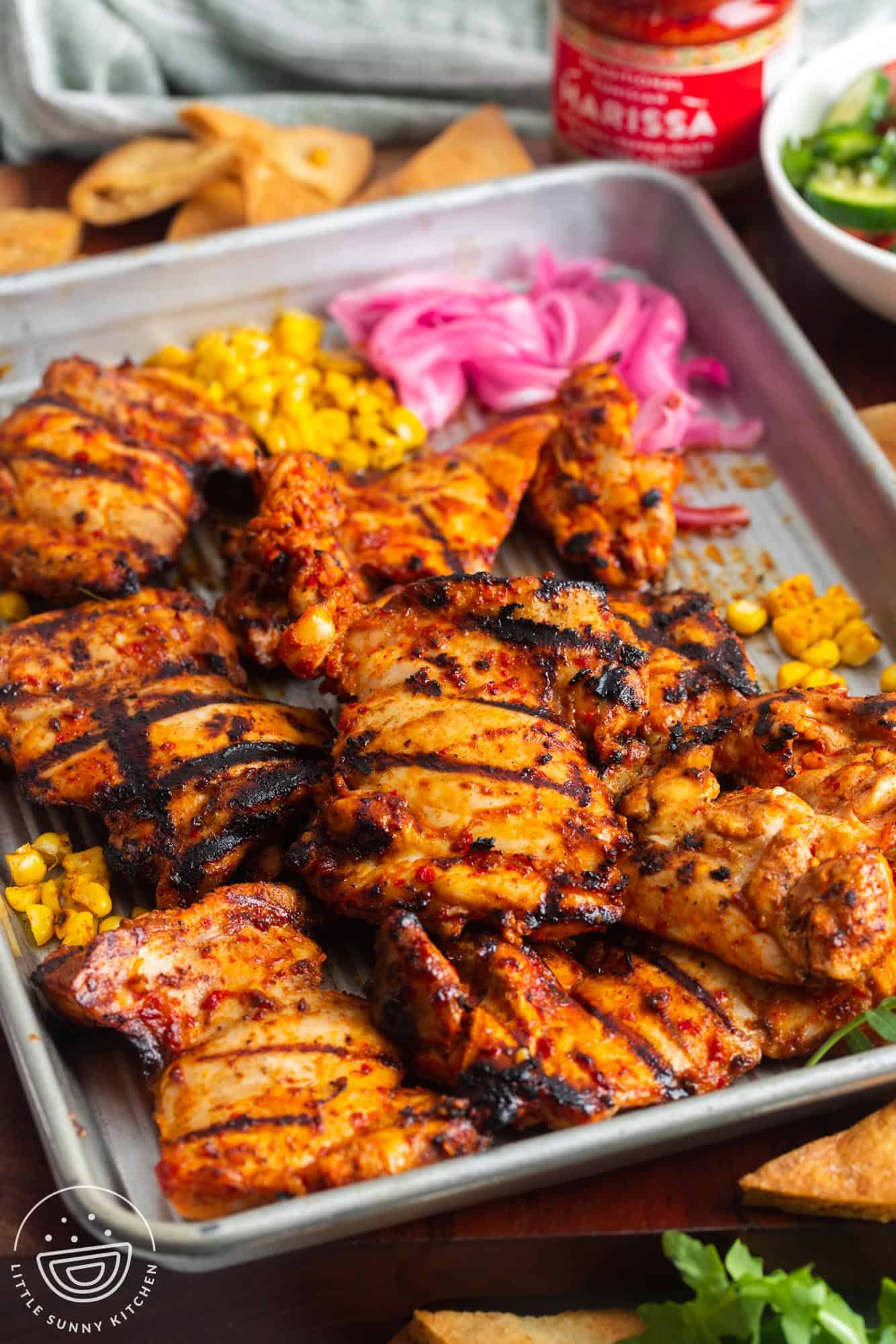 a sheet pan of grilled harissa chicken with onion and corn on the side.