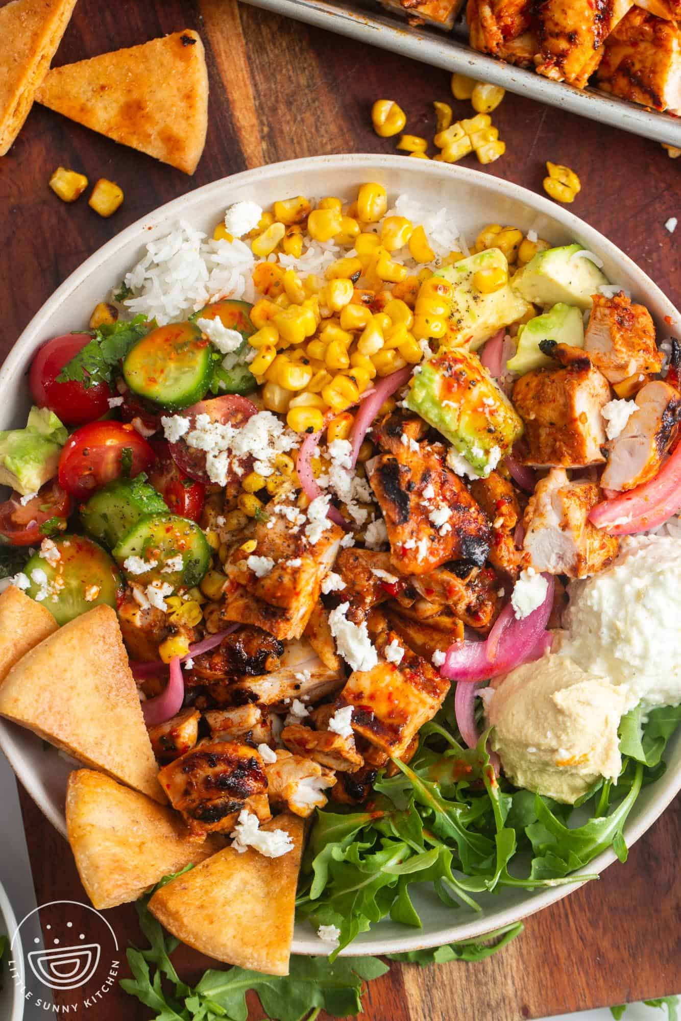 a rice bowl topped with veggies, hummus, pita chips, and grilled harissa chicken.