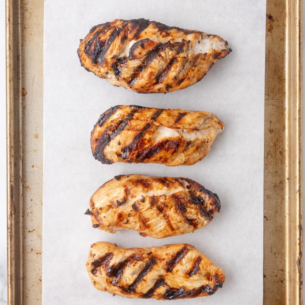 four grilled chicken breasts on a parchment lined baking sheet.